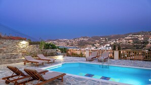 A pool area with lounge chairs, stone flooring, and a view of a hillside town at dusk. - Villa Ariadni Agia pelagia close to the beach up to7-8 quests (Agia Pelagia)