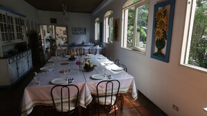 A dining area with a long table set for a meal, surrounded by chairs, with a view of greenery outside through large windows. - Centro Paulus (São Paulo)