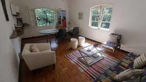 A living room with a sofa, a coffee table, a dining table with chairs, and a large window with a view of trees. - Centro Paulus (São Paulo)