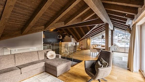 Un loft espacioso con techo alto y vigas de madera, que presenta una sala de estar moderna con sofá, sillón y vistas a un paisaje urbano a través de amplias ventanas.