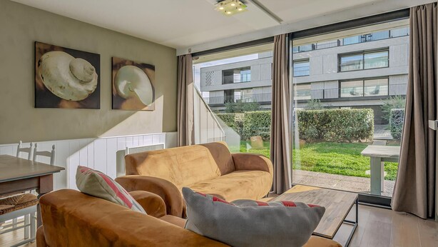 A modern living room with a sofa set, a dining table, and a view of a building and garden through large windows.