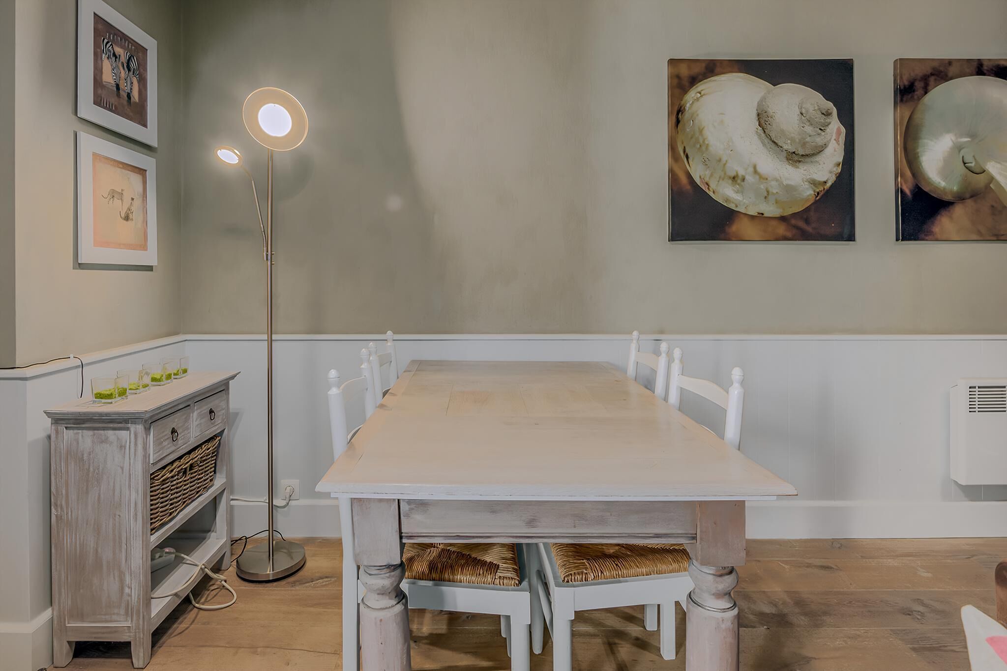 A dining area with a wooden table, white chairs, a floor lamp, and wall art.