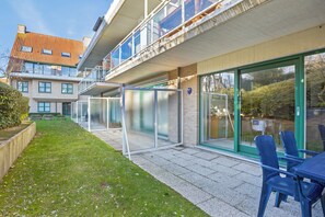Un edificio moderno con ingresso tramite porta in vetro, una piccola area esterna con sedute blu e un prato curato.
