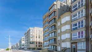 Uma fileira de edifícios residenciais com varandas, céu claro e rua pavimentada.
