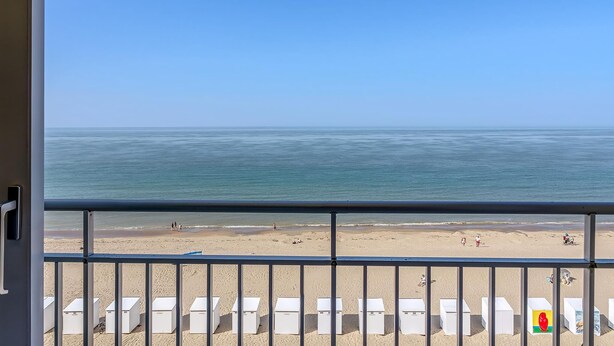 Vista da praia a partir de uma varanda, com espreguiçadeiras brancas e céu claro.