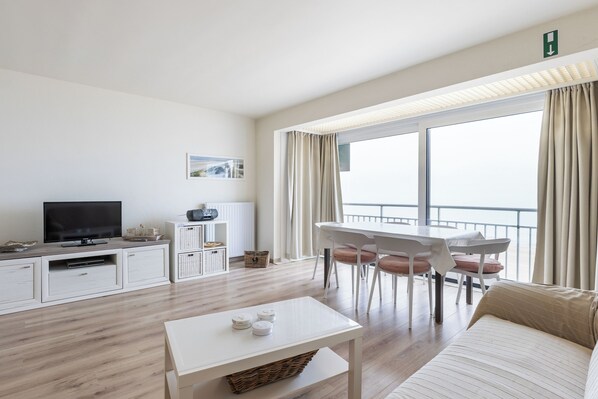 A modern living room with a flat-screen TV, a dining table with chairs, a sofa, and a large window with curtains.