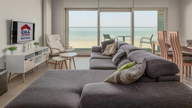 Ein modernes Wohnzimmer mit einem grauen Sofa, einem kleinen Tisch und einem Fernseher auf einem weißen Schrank. Ein großes Fenster bietet Blick auf den Strand.