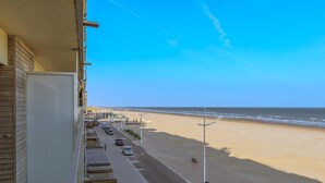 Blick auf einen Strand vom Balkon aus, mit blauem Himmel, Sandstrand und geparkten Autos am Straßenrand.