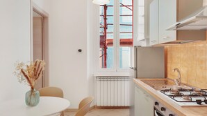 A modern kitchen with a gas stove, oven, and white cabinets. There is a dining table with chairs and a vase of dried flowers. A window with a view of a red building is visible.