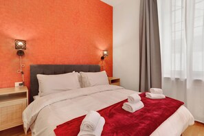Habitación de hotel con una cama grande, dos mesitas de noche, una manta roja y dos lámparas de pared.