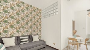 A modern living room with a grey sectional sofa, a dining table with chairs, and a patterned wallpaper.
