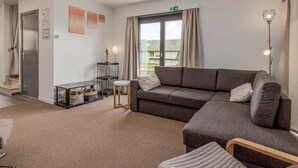 A modern living room with a grey sofa, a small round table, and a floor lamp. There is a door with a handle and a window with curtains. - Peter Pan 97/0201 + G* / (Sint Idesbald)