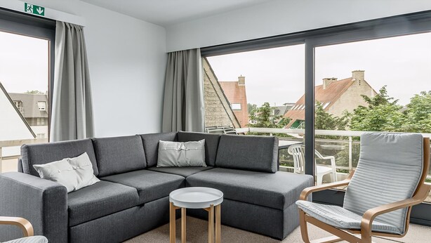 A modern living room with a grey sectional sofa, a round wooden side table, and a grey cushioned chair. - Peter Pan 97/0201 + G* / (Sint Idesbald)