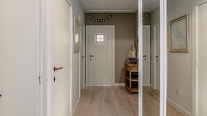 Um corredor com piso de madeira, paredes brancas e uma porta com uma pequena janela. Há uma guirlanda decorativa na parede e uma mesinha com um modelo de navio.