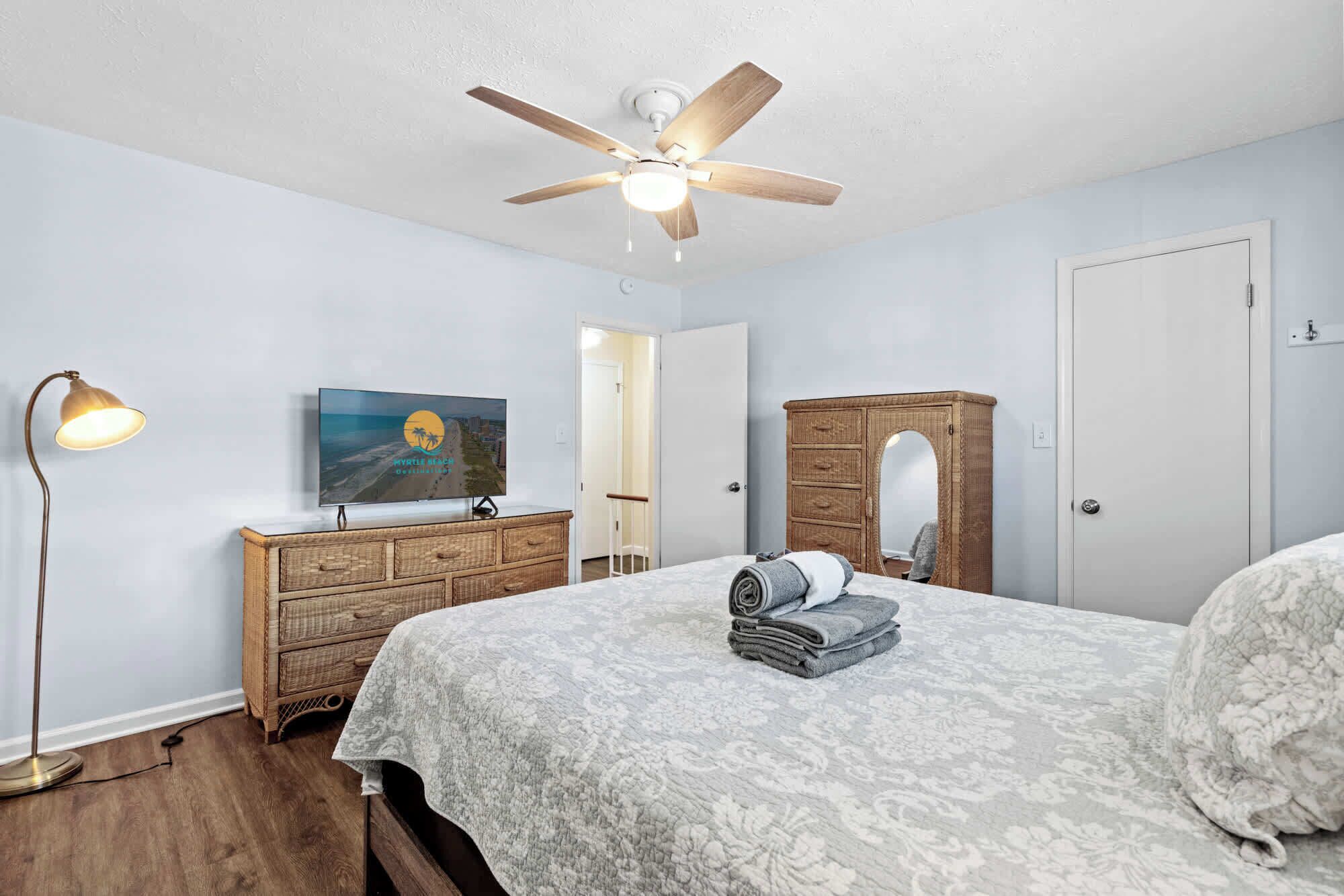 Une chambre à coucher comprenant un lit, un ventilateur de plafond, une commode, un miroir, une lampe et un téléviseur diffusant une scène de plage.