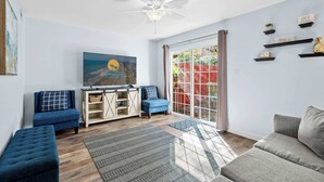 A living room with a sofa, a blue chair, a wooden cabinet, a ceiling fan, and a large window with a view of the outdoors. - Porcher Villas 3 - Near Ocean! (Myrtle Beach)