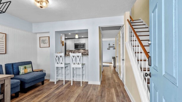 A modern living room with a blue sofa, white chairs, and a wooden dining table. A kitchen is visible through an open doorway, featuring white cabinets and a microwave. A staircase with wooden steps and a blue door leads to another level. - Porcher Villas 3 - Near Ocean! (Myrtle Beach)
