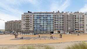 Un immeuble résidentiel situé en bord de mer, doté de nombreux balcons, avec une plage de sable devant et un ciel dégagé.