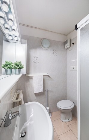 Ein Badezimmer mit Waschbecken, Toilette und einem beleuchteten Spiegel. Auf der Ablage stehen Blumentöpfe.