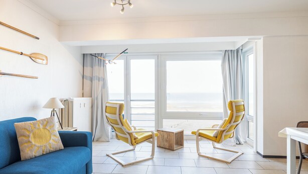 Una sala de estar moderna con un sofá azul, dos sillones amarillos, una mesa de centro de madera y vistas al mar a través de amplios ventanales.
