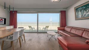 Une chambre d’hôtel moderne avec une table à manger en bois, deux chaises blanches, un canapé en cuir rouge et une grande fenêtre donnant sur la plage.