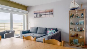 A modern living room with a grey sofa, a wooden dining table, and a glass display cabinet with various collectibles. - 2 bedroom accommodation in Sint Idesbald (Sint Idesbald)