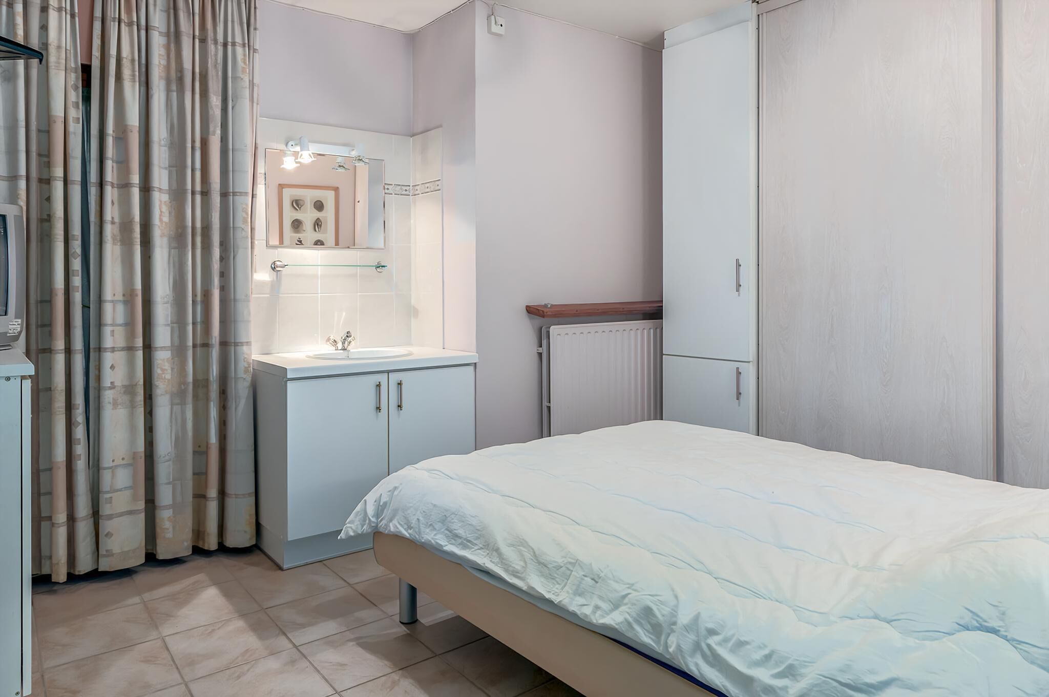A bedroom with a bed, a bedside table, a mirror, a radiator, and a wardrobe.