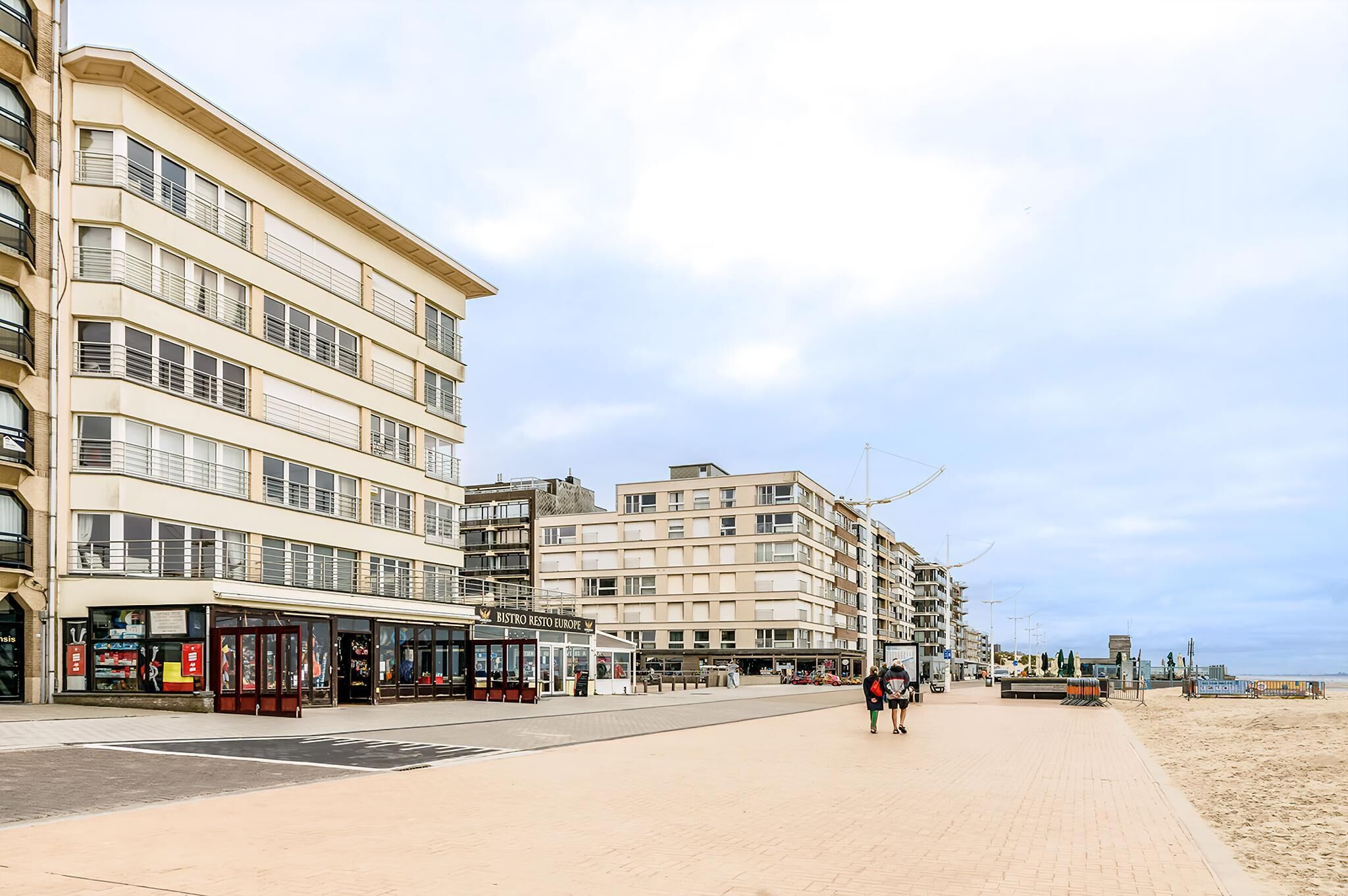 Eine Uferpromenade mit mehrstöckigen Gebäuden, einem Café und einem Strand im Hintergrund.