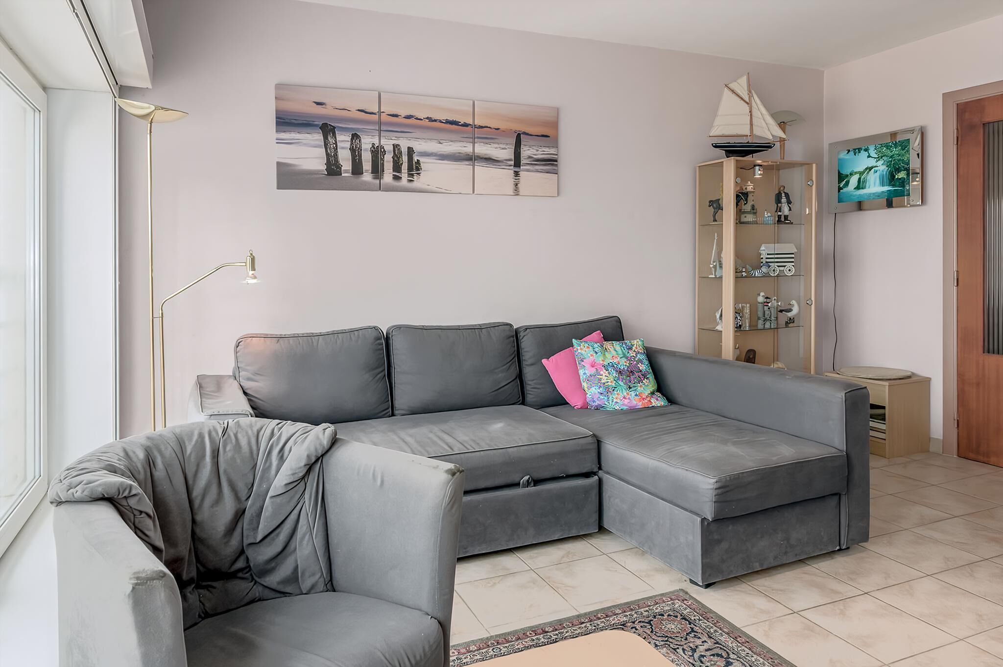 Una sala moderna con un sofá seccional gris, una lámpara de pie, una estantería y un cuadro de una puesta de sol.