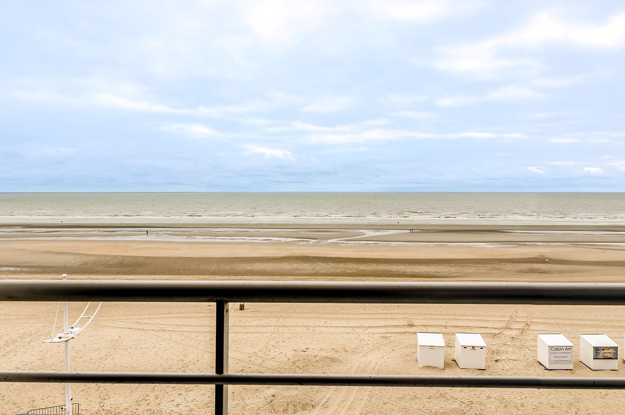 A beach view from a balcony with a railing, featuring a sandy beach, calm sea, and a clear sky.