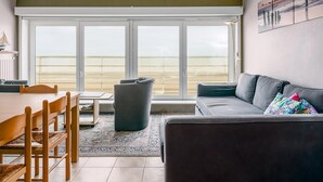 A living room with a large sliding glass door leading to a balcony, a dark grey sofa, a wooden dining table with chairs, and a patterned rug. - 2 bedroom accommodation in Sint Idesbald (Sint Idesbald)