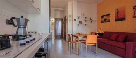 A modern kitchen with a coffee maker, a dining area with a red sofa, and a hallway leading to a bedroom.