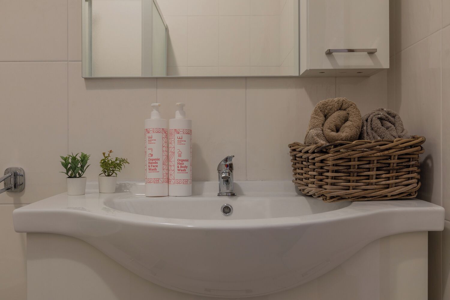 A bathroom sink with two bottles of organic hand and body wash, a small potted plant, a wicker basket with towels, and a mirror above.