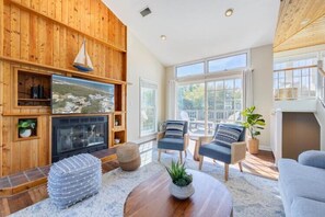 A cozy living room with a fireplace, comfortable seating, and a large window offering a view. - Updated Home, Very Close to the Beach! (Duck)