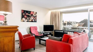 A modern living room with a red sofa, two red armchairs, a wooden coffee table, a TV, and a large window with a view of buildings. - 3 bedroom accommodation in Sint Idesbald (Sint Idesbald)