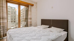 Quarto com uma cama grande, cabeceira escura e vista para uma varanda com cadeiras e um edifício ao fundo.