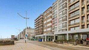 Um edifício moderno de vários andares com varandas, uma área de estar coberta ao ar livre e um céu claro.