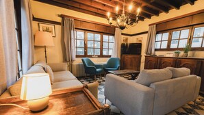 Un salón acogedor con un sofá, un sillón y una mesa de centro de madera con una lámpara. Hay ventanas con cortinas, una araña y un televisor.