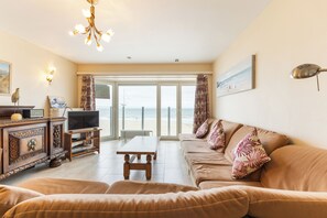 Una sala de estar con sofá, una mesa de centro, televisor y vistas a la playa a través de grandes ventanales.