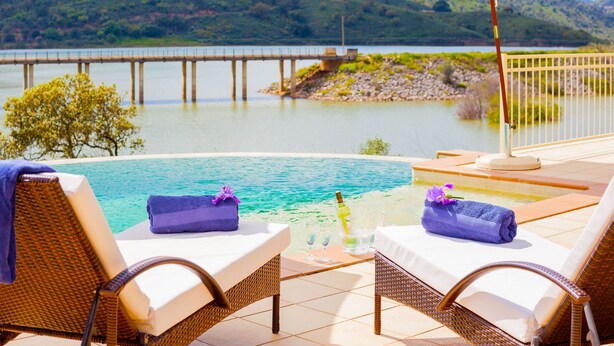A poolside area with lounge chairs, a table with a bottle and glasses, and a view of a lake and bridge. - Casa Dos Amigos in S o Bartolomeu de Messines (Silves)