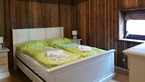 Un dormitorio con techo de madera, una cama con ropa de cama de flores verdes, una mesita de noche con lámpara y una ventana con cortina.