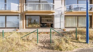 A two-story building with balconies, large windows, and a sandy ground area.