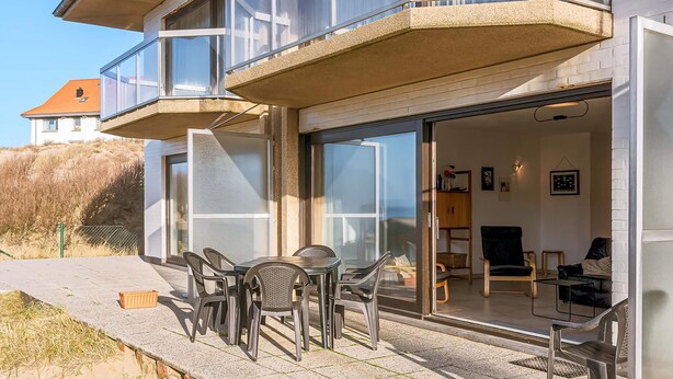 A two-story house with a balcony, a patio with outdoor furniture, and a sliding glass door leading to an interior with a sofa and a table.