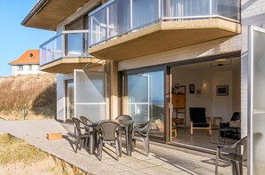 Ein zweistöckiges Haus mit Balkon, einer Terrasse mit Gartenmöbeln und einer Glastür, die zu einem Innenraum mit Sofa und Tisch führt.