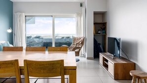 Un salon moderne doté d’une grande table à manger en bois, de sièges confortables, d’un téléviseur à écran plat et offrant une vue sur l’océan grâce à de grandes fenêtres.