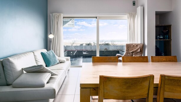 Un salon moderne avec un canapé blanc, une table à manger en bois et des chaises. On y trouve une grande fenêtre avec des rideaux, une bibliothèque et une vue sur l’extérieur.