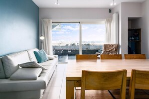 Ein modernes Wohnzimmer mit einem weißen Sofa, einem Holzesstisch und Stühlen. Ein großes Fenster mit Vorhängen, ein Bücherregal und ein Blick ins Freie.