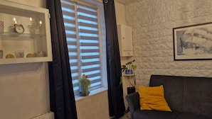 Un salón con sofá gris, cortina negra, ventana con persianas, estante empotrado con objetos decorativos y un cuadro en la pared.