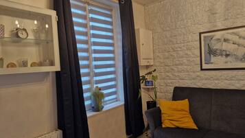 Un salon avec un canapé gris, un rideau noir, une fenêtre avec des stores, une étagère fixée au mur ornée d’objets décoratifs et un tableau encadré accroché au mur.