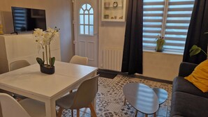 Un comedor con una mesa blanca, sillas y una orquídea en maceta. A la izquierda, hay un televisor sobre un mueble.
